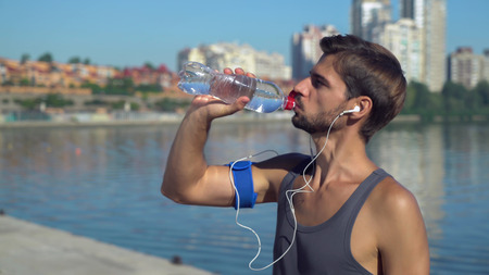 Young athletic guy in shirt after run standing near the river or sea, drinking water from a bottle and speaking.Mixed race happy smiling man rest after workout in the city with urban view. Healthy sport life concept.の写真素材