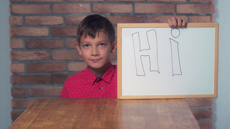 child climbs under the desk holding flipchart with lettering hi the background red brick wall. Schoolboy shows hand gesture hello. Preadolescent wearing in casual red shirt.の写真素材