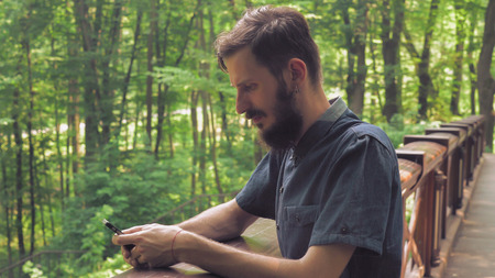 A young man using mobile phone wearing in a blue shirt with a beard standing on the bridge in the...