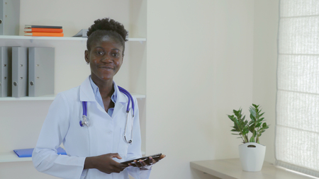 Young doctor standing with happy candid smile in hospital. Afro american woman wearing medical uniform white coat and phonendoscope holding digital tablet. Smiling professional staff in clinic.の写真素材
