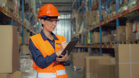 Manager female working at warehouse. Attractive young woman worker wearing uniform hard hat and orange vest, counting box for delivery filling up form holding pen.の写真素材