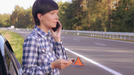Young woman standing on the driveway near broken car holding card and mobile calling local garage or mechanic. Caucasian young female talking by smartphone on the roadside.の写真素材