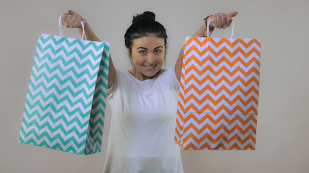 happy beautiful caucasian woman with black hair showing colored paper bags with purchases. Portrait young girl wearing casual white t-shirtの写真素材