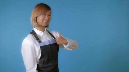 attractive korean woman with blond hair wearing white shirt and apron looking at the camera smiling. portrait young asian girl posing showing sign like on blue background in studioの写真素材