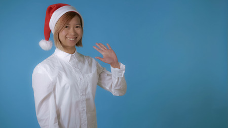 portrait young asian female posing wearing christmas hat waving hand say hello on blue background in studio. attractive korean woman with blond hair wearing white casual shirt looking at the camera smilingの写真素材