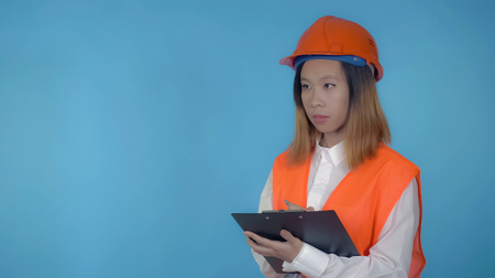 young asian woman manager in logistic company filling form for delivery on blue background in studio . attractive millennial girl wearing white casual shirt and orange hard hat and vest looking at the camera with cheerful happy smileの写真素材