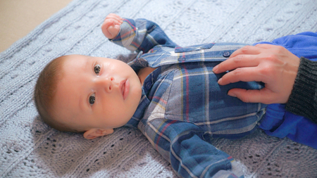 Little baby boy lying on the bed holding mother hand. Cute child wearing blue shirt.