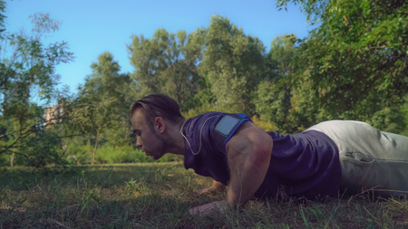 bodybuilder trains in park with green grass and trees. Caucasian adult man push-up outdoors.