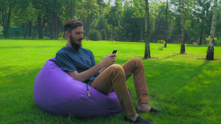 A young guy with a beard in a blue shirt sits on a chair in the park pufi and gaining a message on your phone, smiling.の写真素材