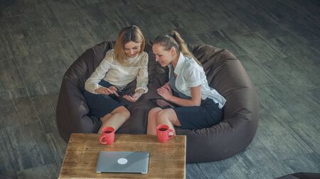 Two attractive business woman relaxing on a sofa and watching photo on the phone. Smile and discuss photos, pleasing appearance. Static videoの写真素材
