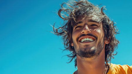 A young man stands outdoors, smiling widely with the wind tousling his hair. The bright blue sky and sunny weather create a cheerful atmosphere, perfect for a day at the beach.の素材