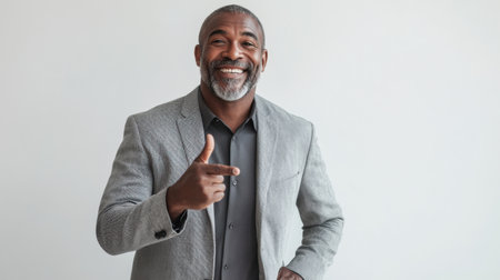 A middle-aged man with dark skin wears a gray suit and smiles confidently while pointing. He stands in an indoor setting with a neutral background, exuding positivity and charm.の素材