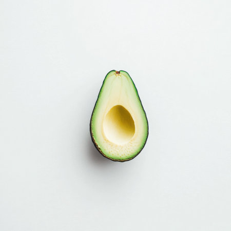 A fresh green avocado is sliced in half, showing its creamy yellow-green flesh and surrounding dark green skin. The avocado rests on a light-colored surface, emphasizing its natural color.の素材