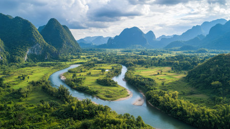 A river gracefully curves through a vibrant green valley, surrounded by imposing mountains under a partly cloudy sky. The landscape showcases nature's beauty.の素材