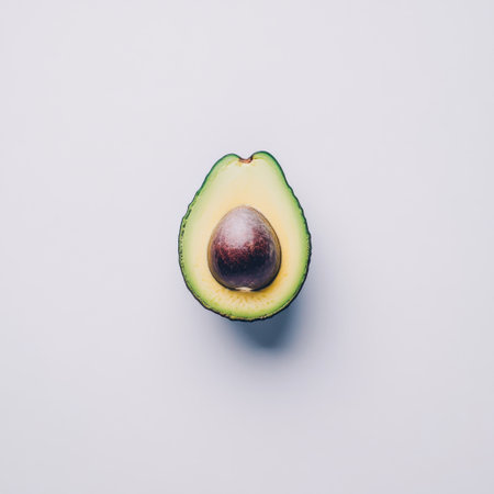 A ripe avocado is cut in half, displaying its green flesh and large brown seed. This simple setup highlights the fruit's smooth texture against a clean white surface.の素材