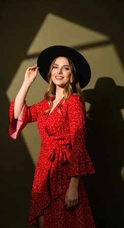 A young woman stands confidently wearing a vibrant red dress with bell sleeves and a wide-brimmed hat. Her playful pose highlights the interesting shadow patterns on the wall behind her.の素材