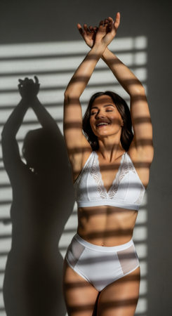 A woman stands gracefully in white lingerie, stretching her arms overhead while sunlight casts playful shadows across her body. The setting feels warm and inviting.の素材