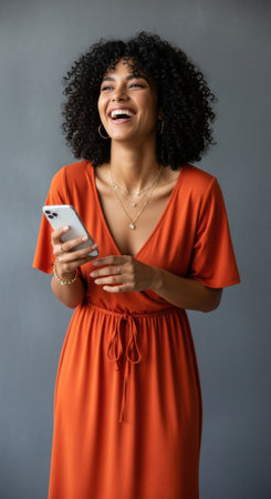 A joyful woman with curly hair wears a flowing orange dress and holds a phone while standing against a gray wall. She is smiling bright, radiating positivity.の素材
