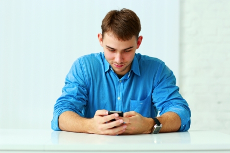 Young happy casual businessman sitting at the table and typing on smartphone in officeの写真素材
