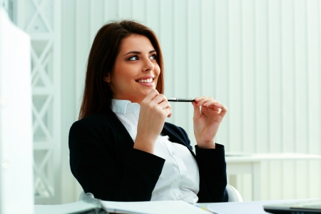 Young smiling businesswoman sitting at the table and looking away at copyspace in officeの写真素材