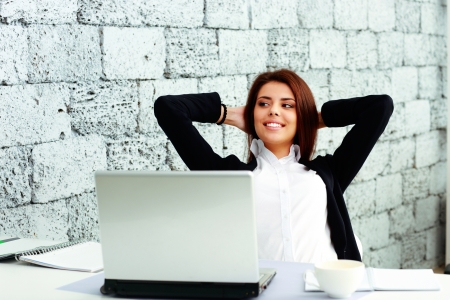 Young happy businesswoman sitting at the table in office near brick wallの写真素材