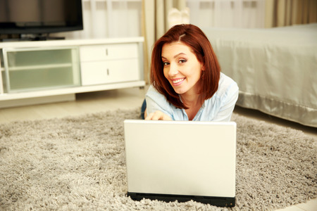Smiling woman lying on the carpet with laptop at homeの写真素材
