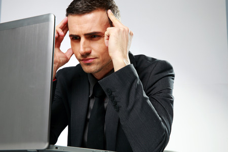 Thoughtful businessman sitting at the table with laptop on gray backgorund