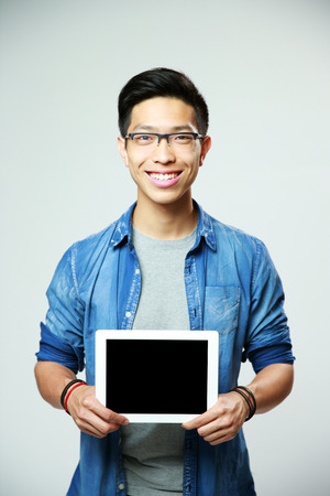 Young cheerful asian man showing tablet computer screen on gray backgroundの写真素材