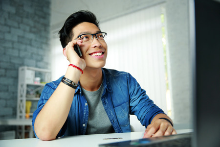 Young happy asian man talking on the phone at his workplace in office