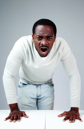 Portrait of a angry african man shouting and leaning at the table over gray backgroundの写真素材