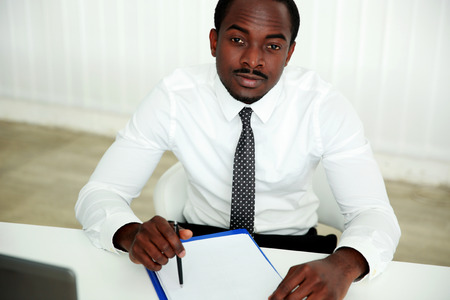 Pensive african man sitting at the table and signing document in officeの写真素材