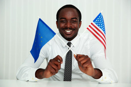 African businessman sitting at the table and holding USA and European flagsの写真素材