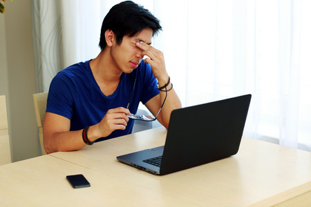 Young tired man sitting at the table with laptop at home