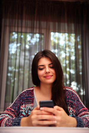 Happy woman sitting at the table and using smartphone at homeの写真素材