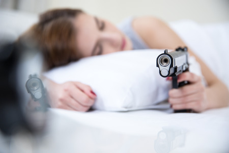 Woman in bed sleeps with hand on gun weapon home security. Focus on gunの写真素材