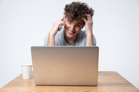 Happy young man sitting at the table with laptop and talking on the phoneの写真素材