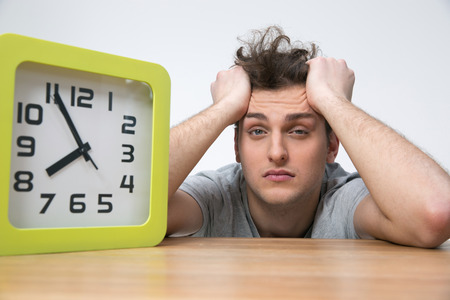Tired young man sitting at the table with big clock