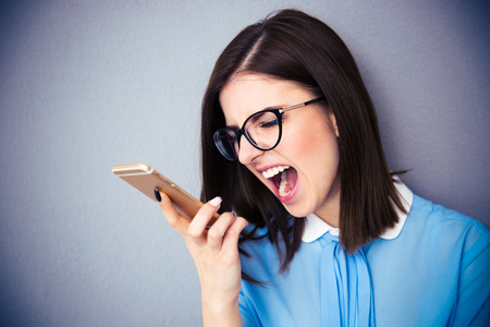 Angry businesswoman shouting on smartphone. Wearing in blue shirt and glasses. Standing over gray...