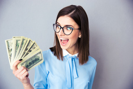 Cheerful businesswoman holding bills of dollar over gray background. Wearing in blue shirt and glasses.の写真素材
