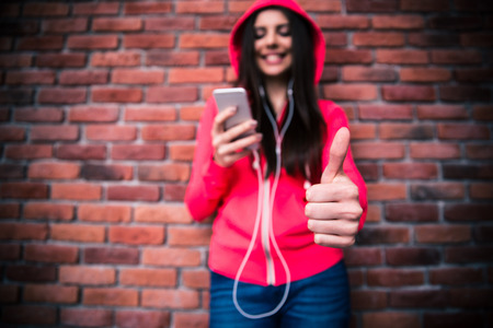 Smiling young woman with smartphone and headphones showing thumb up over brick wall. Focus on hand