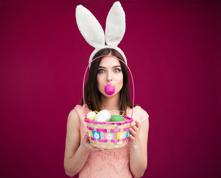 Woman in bunny ears holding Easter egg basket over pink background. Looking at camera. Holding egg...