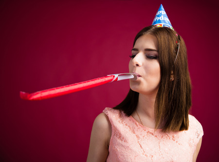 Young woman blowing in party whistle over pink background. Celebrating birthday.の写真素材