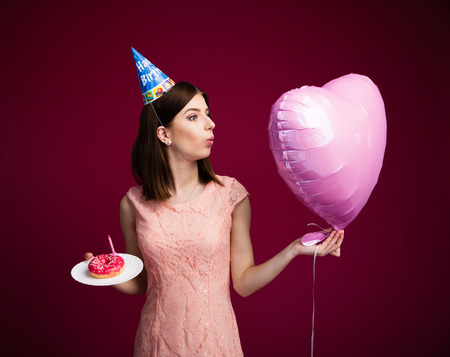 Woman holding heart shaped balloon and cake with candle over pink background. Blowing on balloon