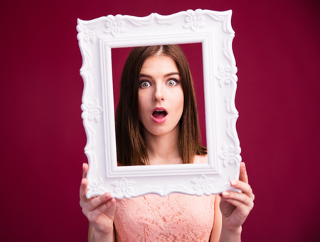Surprised woman looking at camera through frame over pink backgorundの写真素材