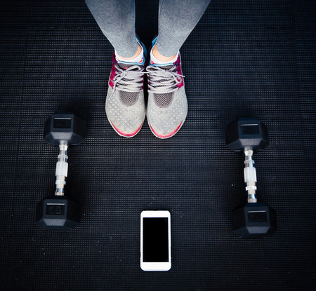 Closeup image of a female legs with dumbbells and smartphone on the floor at gymの写真素材