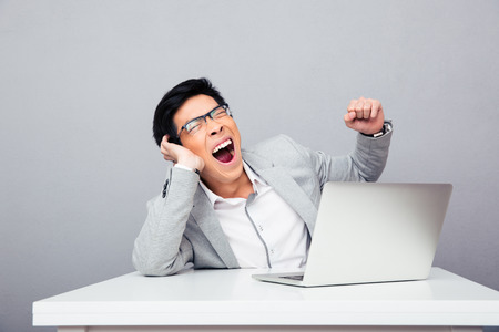 Businessman sitting at the table with laptop and yawning over gray backgroundの写真素材