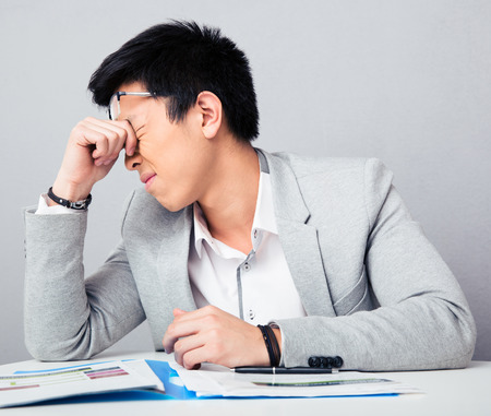 Tired businessman sitting at the table and rubbing his eyes over gray backgroundの写真素材