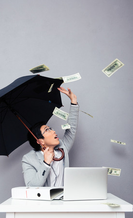 Asian businessman sitting at the table with umbrella with rain of money over gray background. Looking upの写真素材