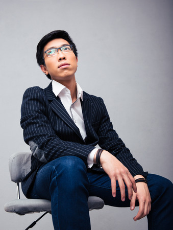 Pensive young businessman sitting on the chair over gray background and looking upの写真素材