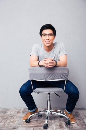 Full length portrait of a cheerful asian man sitting on the chair and looking at cameraの写真素材